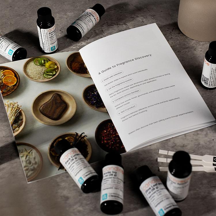 A scattering of 1 oz bottles of fragrance oil, blotter strips, and a booklet on a grey tabletop. 
