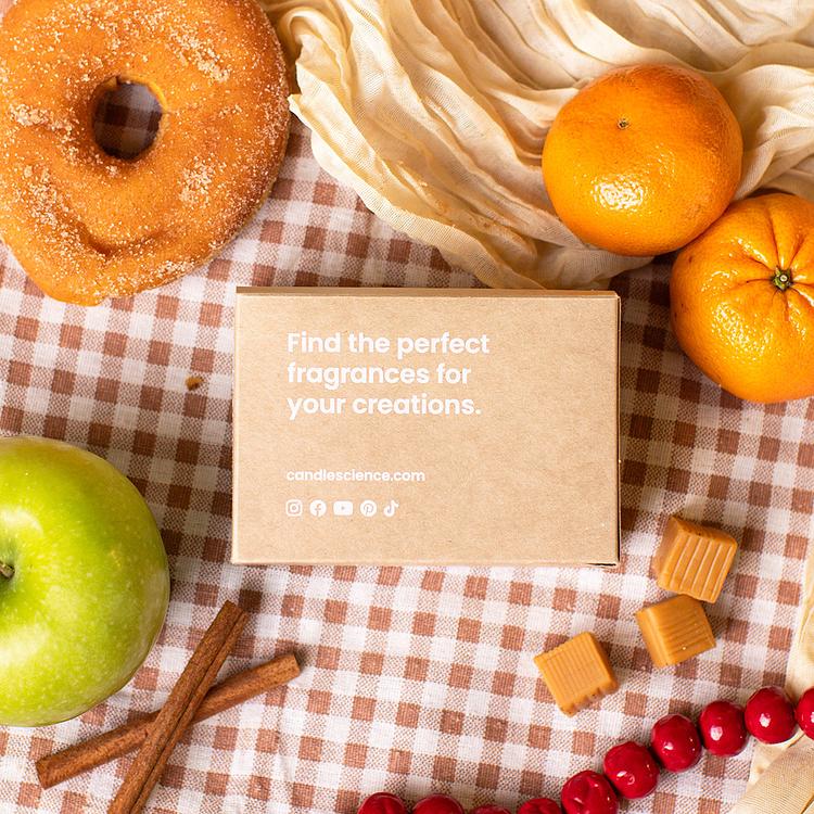 A kraft fragrance flight box on a red and white checkerboard tablecloth background with apples, cinnamon, donuts, and other fragrance note elements