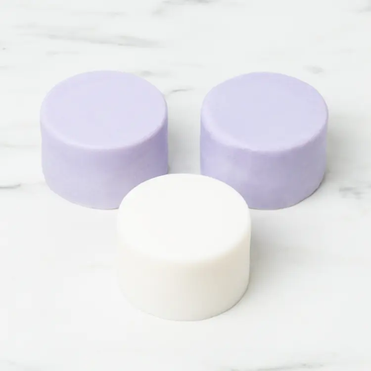 Three shampoo bars on a white marble tabletop. One shampoo bar is white and the other two are lilac.