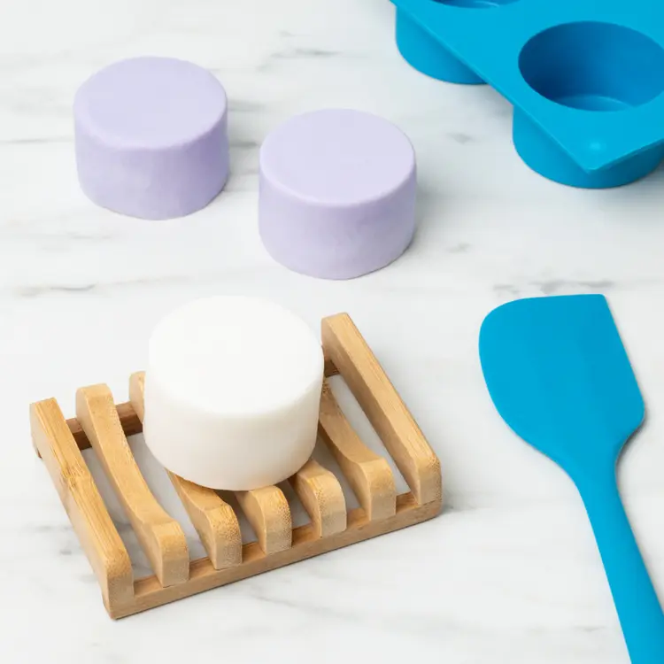 A white, circular shampoo bar on a bamboo soap holder. Two lilac shampoo bars are in the background with a soap mold and a spatula