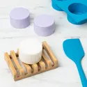 A white, circular shampoo bar on a bamboo soap holder. Two lilac shampoo bars are in the background with a soap mold and a spatula