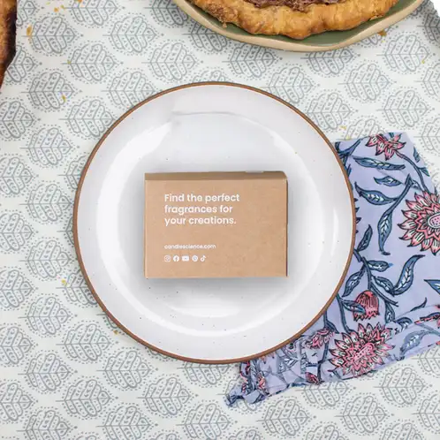A kraft fragrance flight box on a linen tablecloth with a multicolors blue and purple linen napkin tucked underneath a white plate with gold trim.