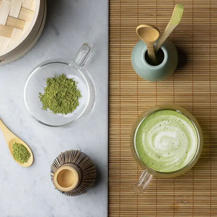 A rattan mat and a white tablecloth showing matcha, bamboo wisks, and hot matcha to represent Mindful Morning Fragrance Oil