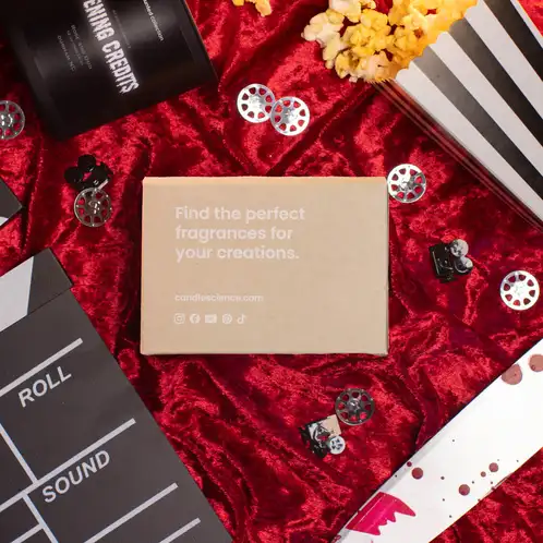 A kraft fragrance flight box that reads "find the perfect fragrances for your creations" on top of red crushed velvet surrounded by movie theater popcorn, film reels, and a prop knife