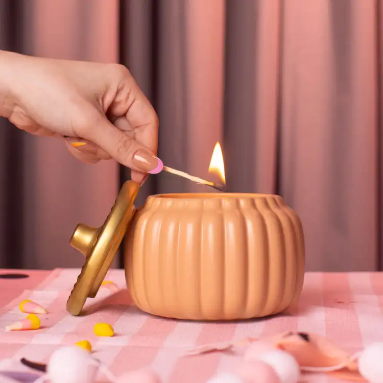 Harvest Orange Ceramic Pumpkin Jar with Gold Matte Lid on its side. A hand and match are in frame lighting the candle on a sweetly spooky pink background