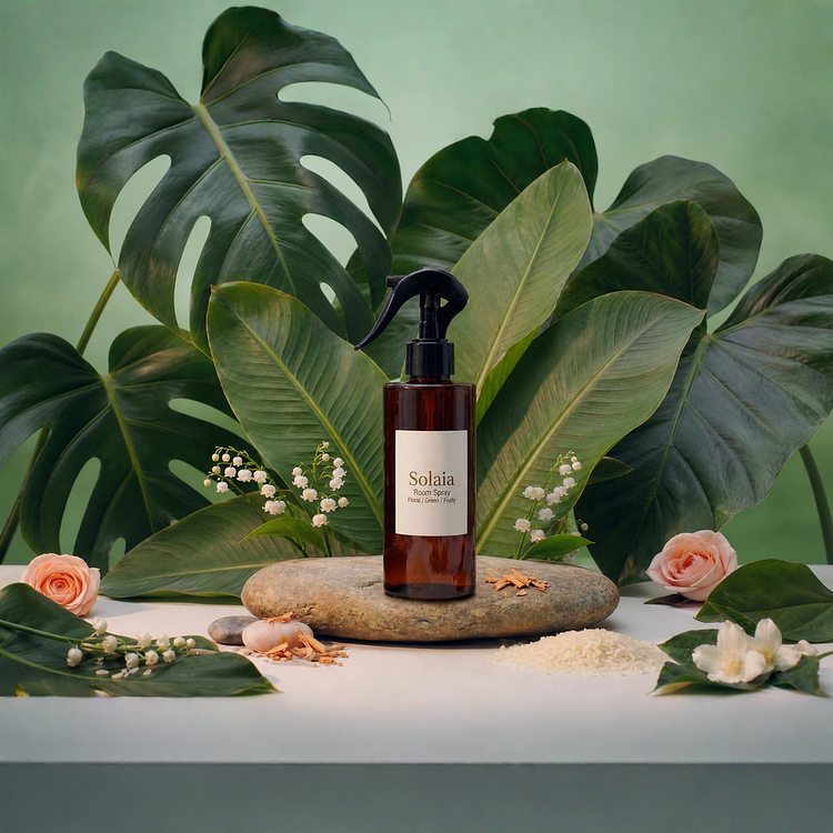 An amber glass bottle of Solaia Room Spray with a black trigger spray top, displayed on a flat river stone surrounded by green tropical leaves, lily of the valley, pink roses, and jasmine.